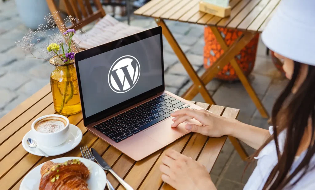 Osoba siedzi przy stoliku w kawiarni na świeżym powietrzu z laptopem wyświetlającym logo WordPress, delektując się kawą i rogalikiem, być może szykując się do aktualizacji strony www lub zarządzania ważną aktualizacją WordPress.