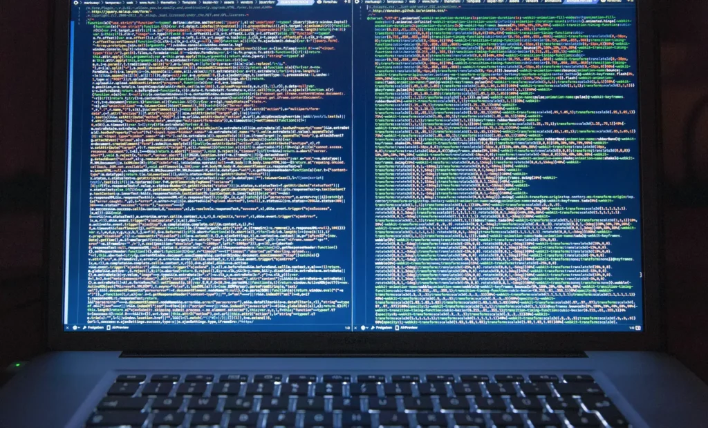 Ekran laptopa wyświetlający linie kolorowego kodu w dwóch oknach obok siebie, sugerujący rozwój oprogramowania lub programowanie — prawdopodobnie skupione na zadaniach takich jak odwirusowywanie WordPressa. Klawiatura jest widoczna na dole obrazu.