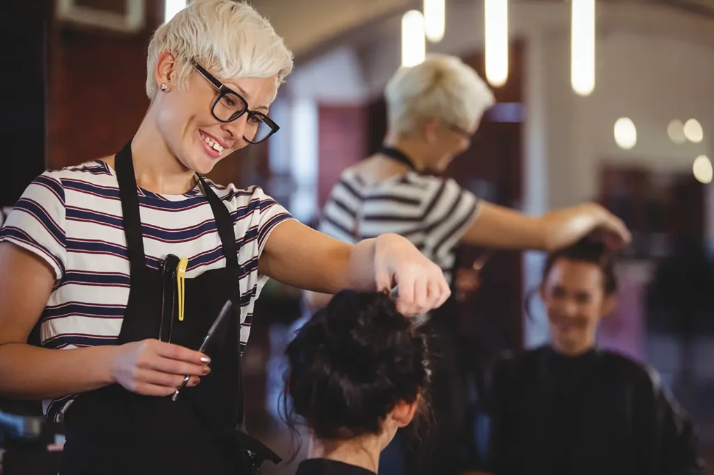 Uśmiechnięta fryzjerka z krótkimi blond włosami i w okularach układa włosy klientce w salonie w Rzeszowie, trzymając nożyczki i grzebień. Klientka siedzi, a oboje odbijają się w lustrze za nimi. Idealne do pozycjonowania wizytówki Google.